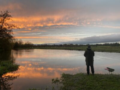 A solitary angler stands with his fishing rod in the sunset on a river