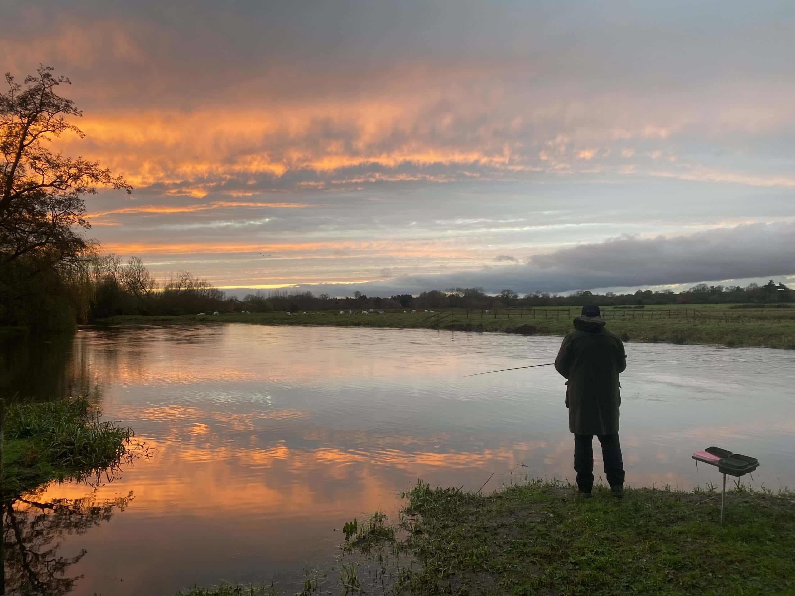 A solitary angler stands with his fishing rod in the sunset on a river